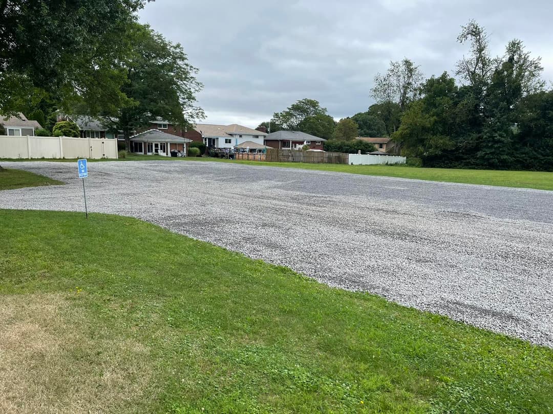 Gravel parking lot in residential area with houses and green trees in the background.