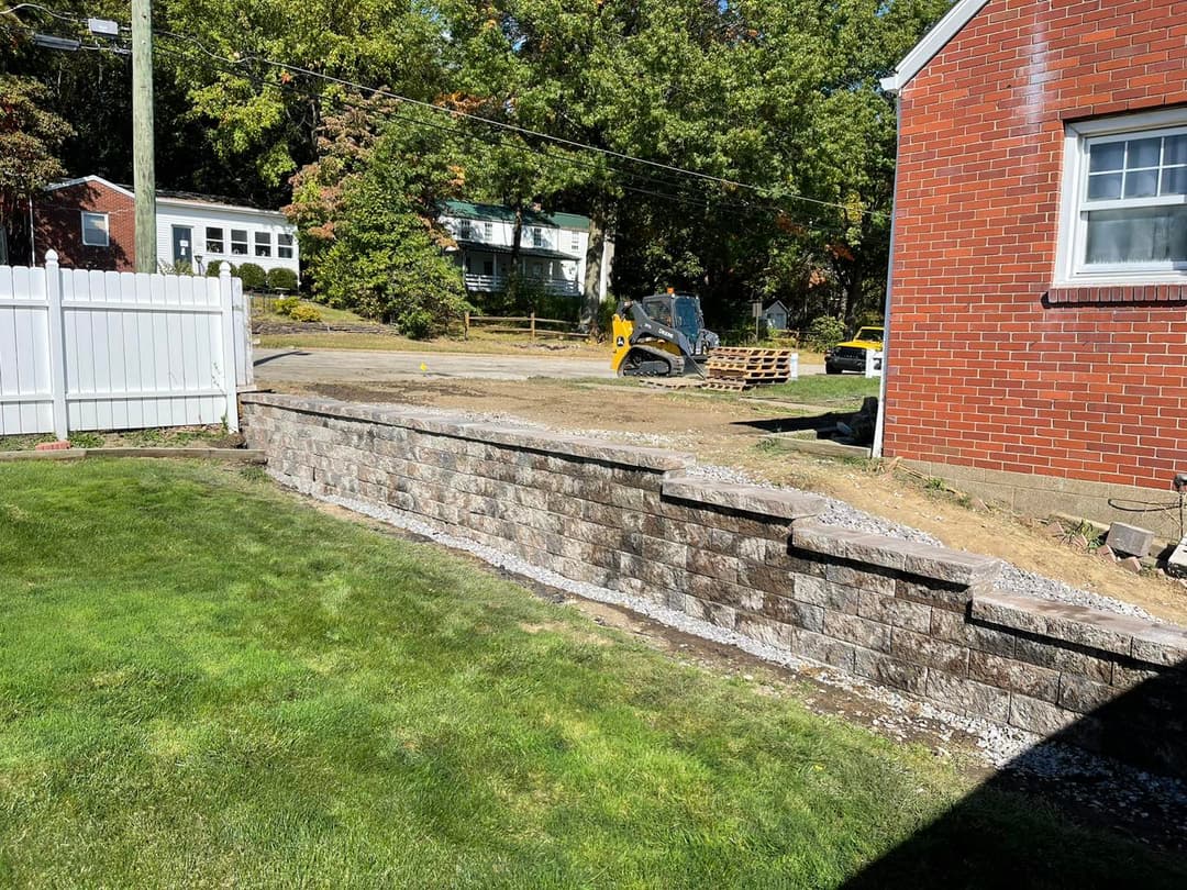 Retaining wall installation in a landscaped yard with machinery and brick house in background.