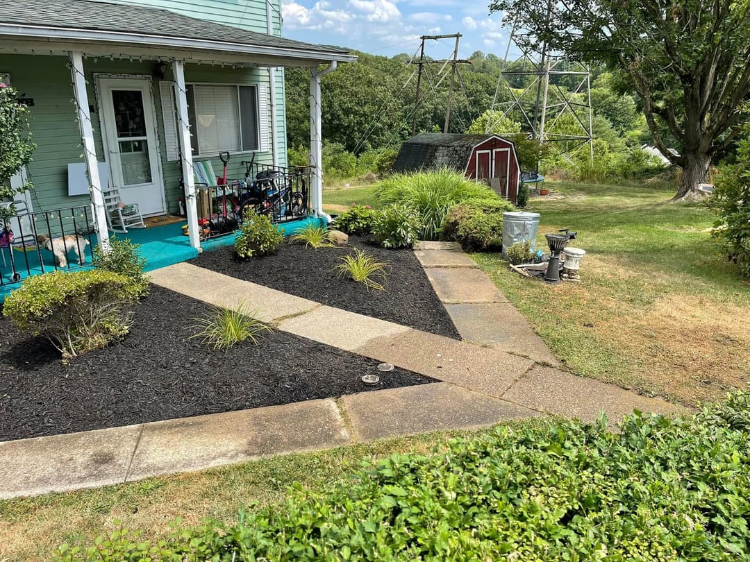 Front yard of a green house featuring a stone pathway, flower beds, and utility shed.