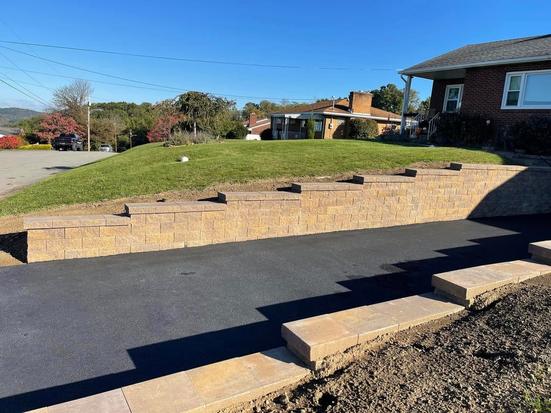 Newly built stone retaining wall alongside paved driveway and green lawn in sunny residential area.