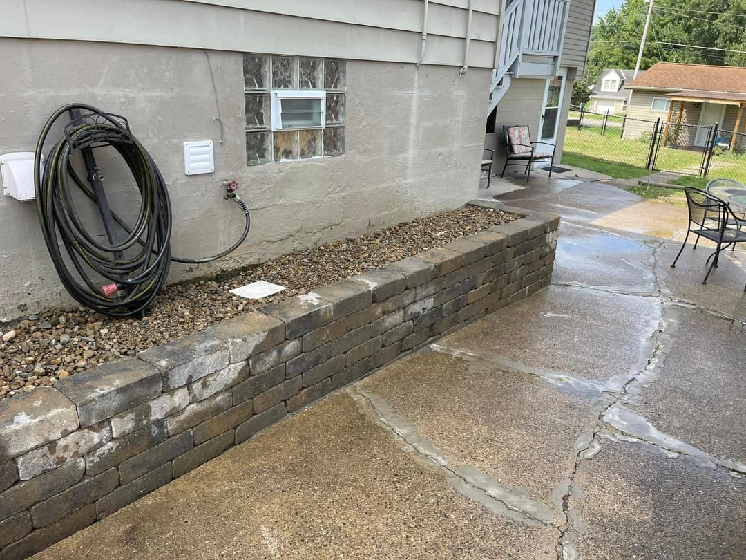 Patio area with stone wall, garden hose, and concrete pavement, featuring clear drainage lines.
