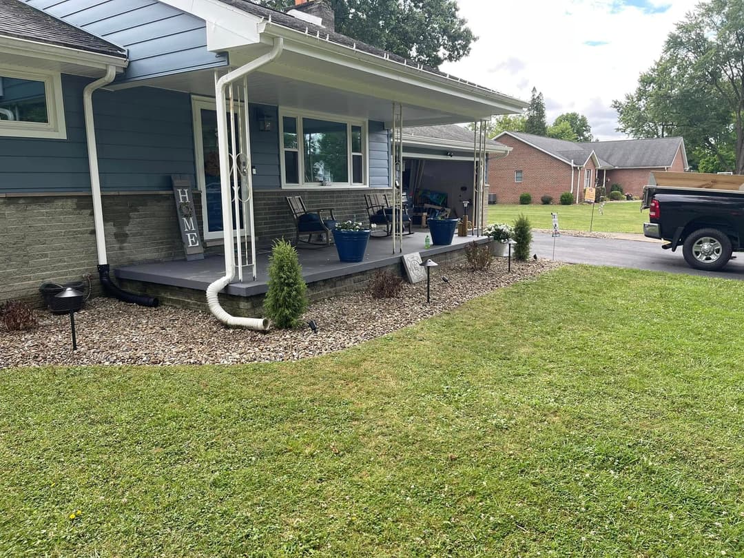 Front porch of a blue house with rocking chairs, potted plants, and green lawn.