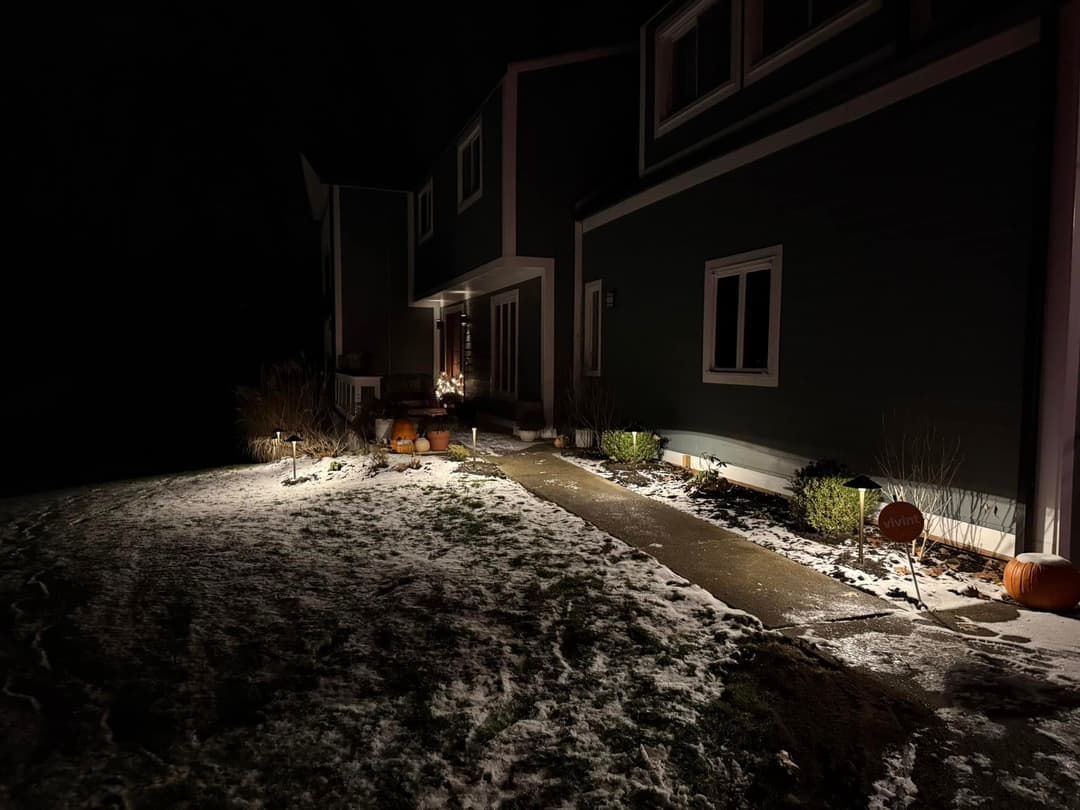 Nighttime view of a house exterior with snow, lit pathway, and seasonal decorations.