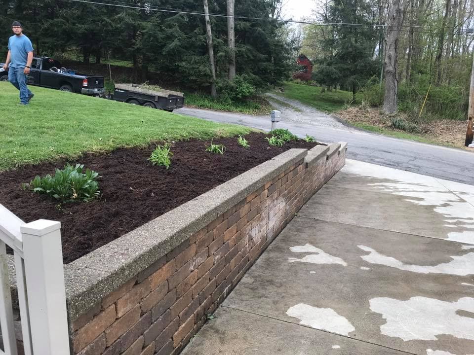 Man gardening near a landscaped stone wall with fresh mulch and plants in a residential area.