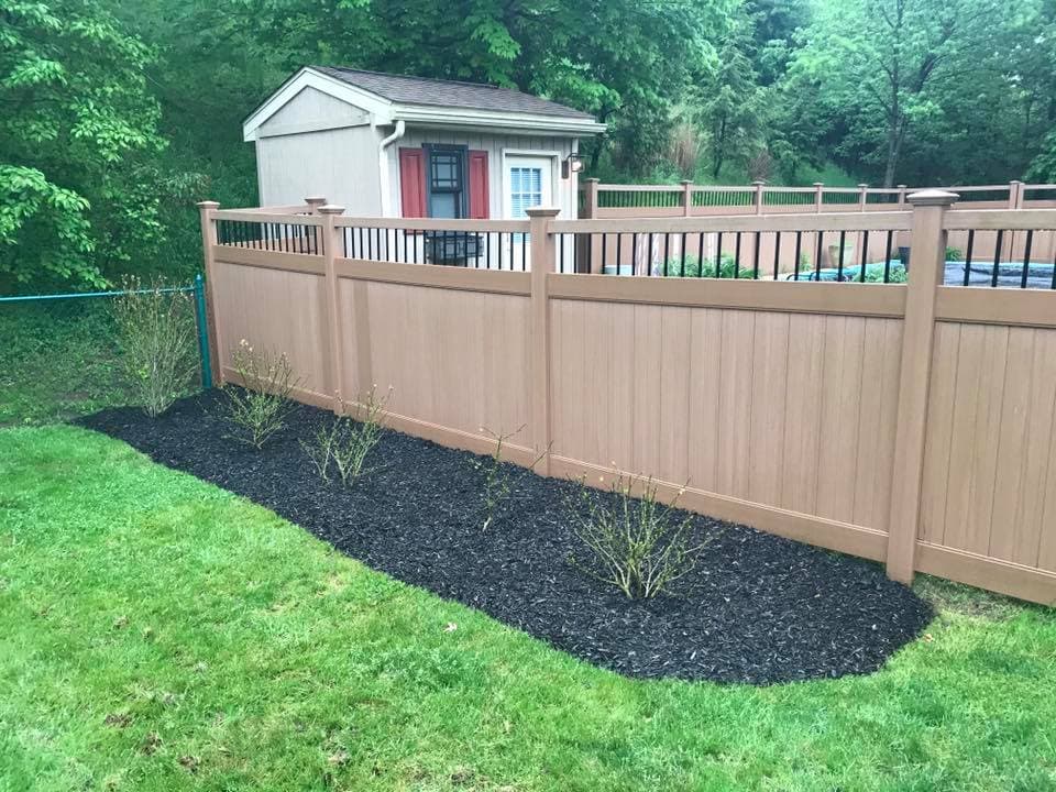 Lawn area with mulch and shrubs near a wooden fence and a garden shed in a green landscape.