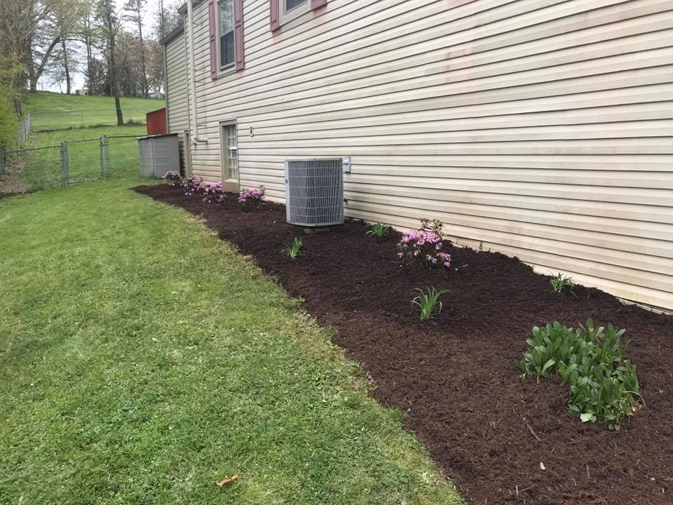 Flower beds with pink blooms and fresh mulch beside a house and air conditioning units.