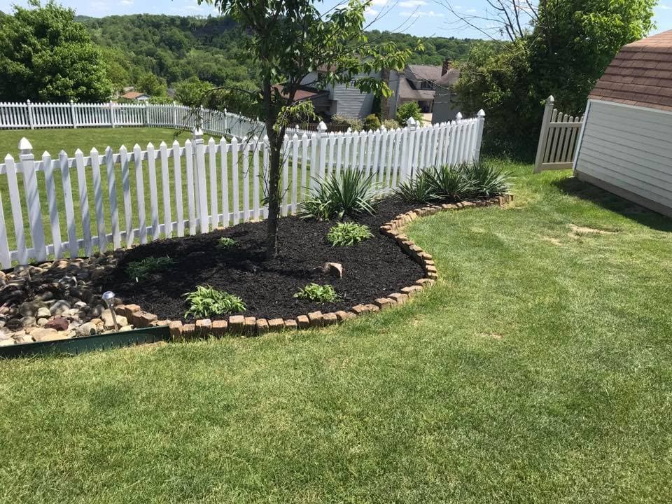 Landscaped garden with a tree, mulch, and decorative stones beside a white picket fence.