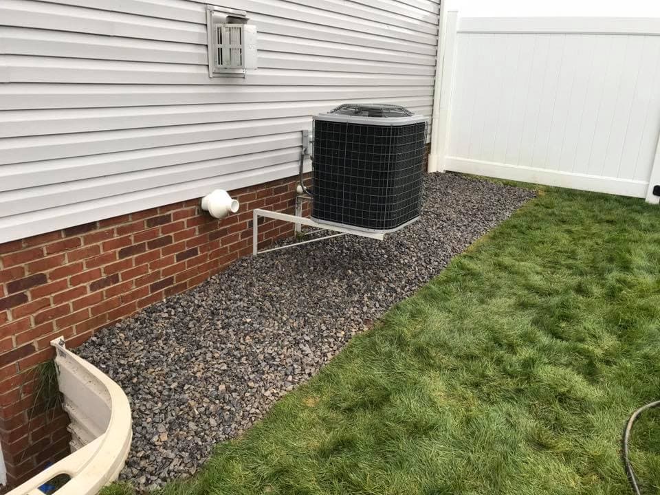 Air conditioning unit on gravel base beside a house with manicured lawn and white fence.