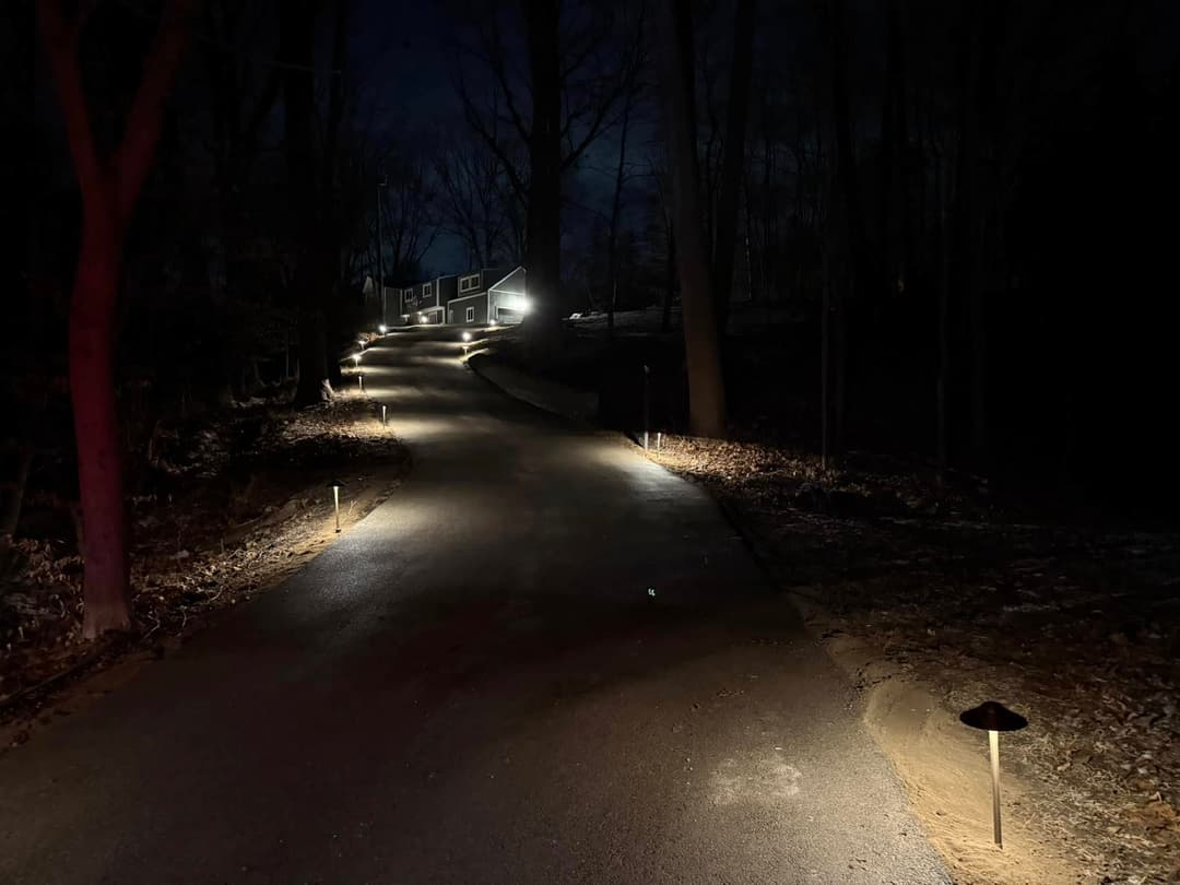 Illuminated driveway at night with pathway lights leading to a house in wooded area.