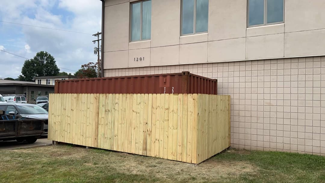 Storage container surrounded by wooden fence next to a building with large windows.