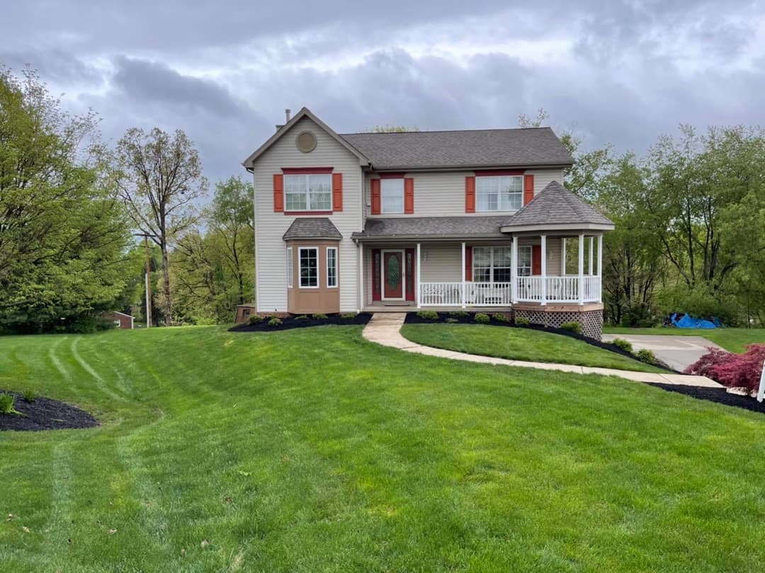 Two-story suburban home with a spacious lawn, porch, and dark green landscaping under a cloudy sky.