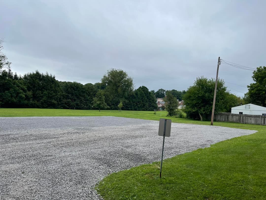 Gravel parking lot surrounded by green grass and trees on a cloudy day.
