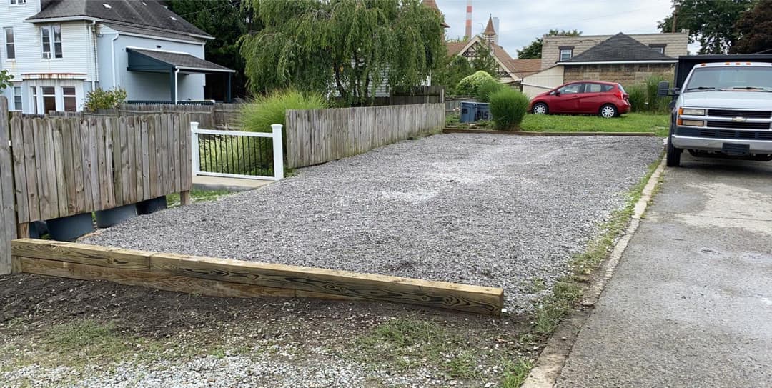 Gravel parking lot next to residential buildings, featuring wooden border and adjacent truck.