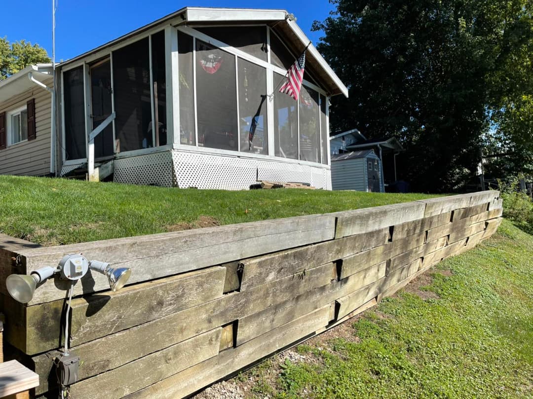 Wooden retaining wall in front of a house with a screened porch and American flag.