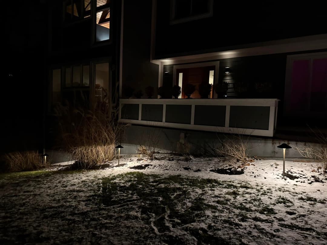 Snow-covered yard at night with house lighting and garden features visible.