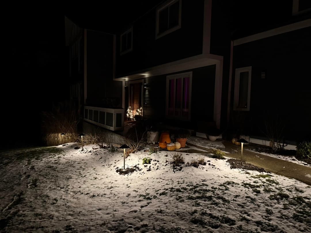 Snow-covered yard at night with illuminated pathway and pumpkins near the entrance.