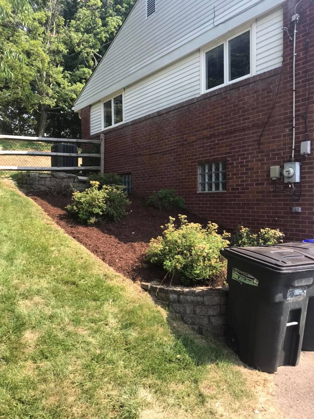 Brick exterior of a house with landscaped garden and trash bin in sunlight.