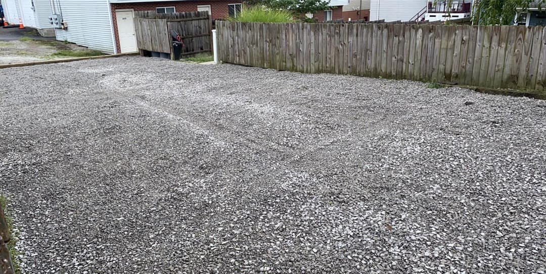 Gravel driveway in empty lot with wooden fence and buildings in background.