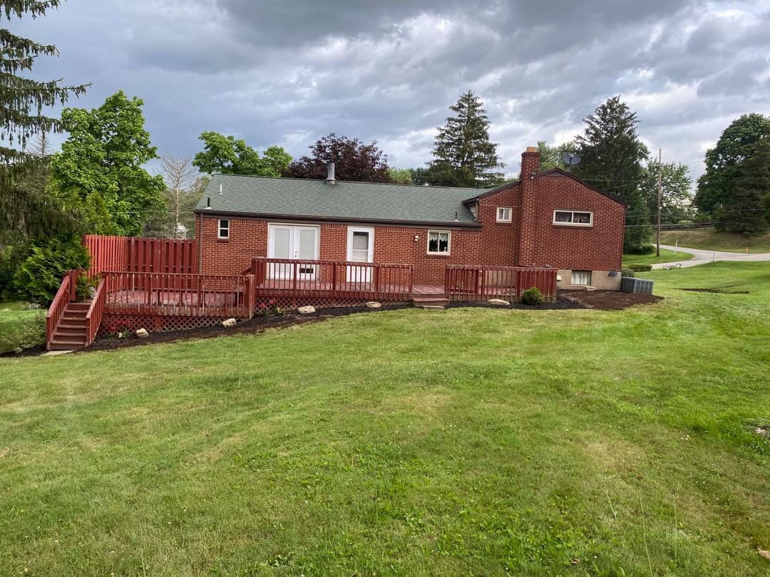 brick house with red deck on green lawn under cloudy sky, surrounded by trees