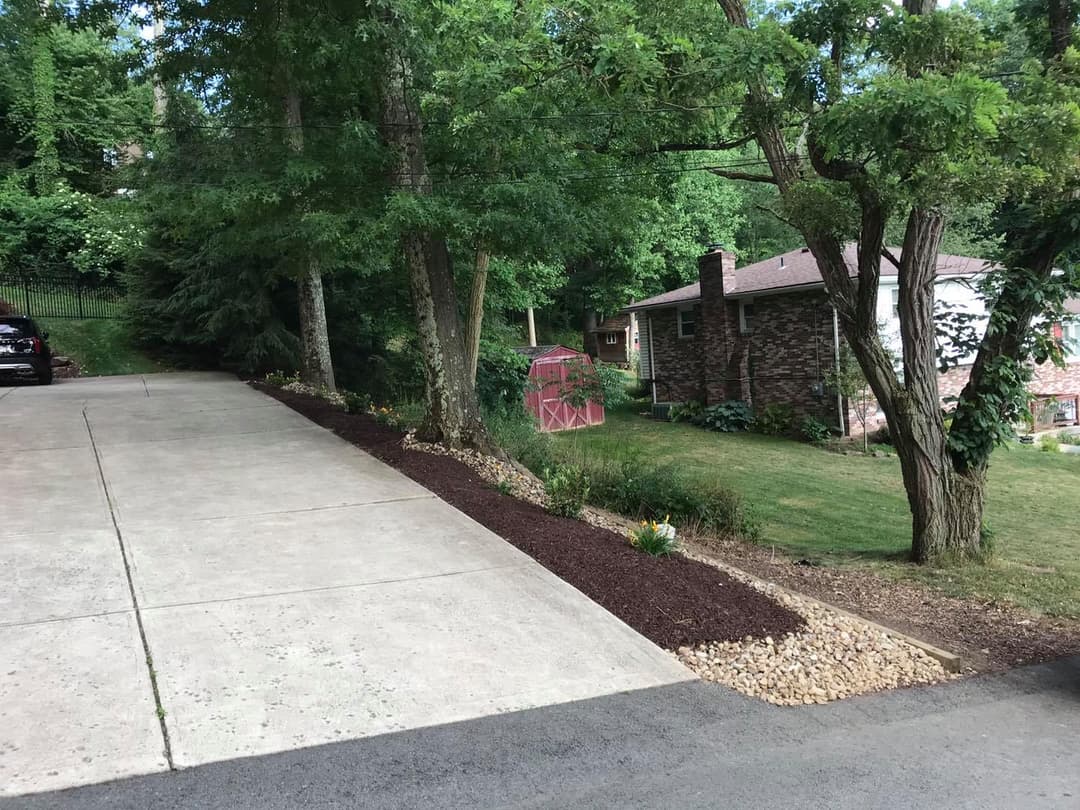 Driveway lined with mulch and landscaping, leading to a brick house in a wooded area.