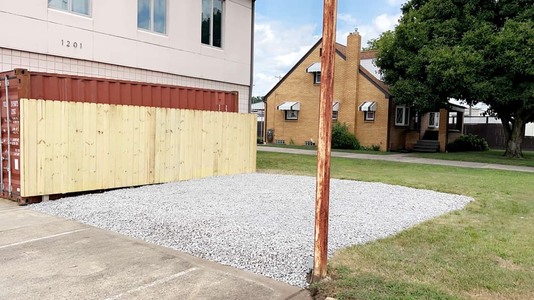 Gravel lot with shipping container and wooden fence, residential house in background.