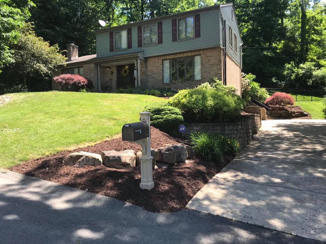 Two-story brick house with landscaped yard and mailbox, surrounded by trees and shrubs.