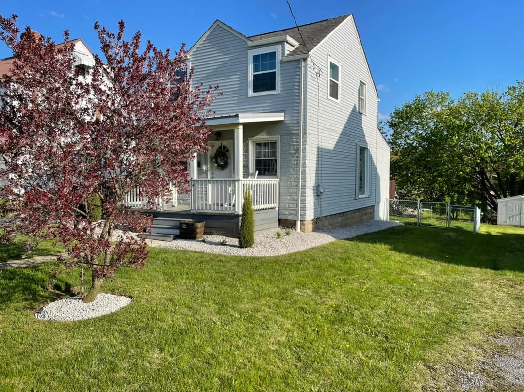 Charming gray house with porch, landscaped yard, and vibrant red tree under blue sky.