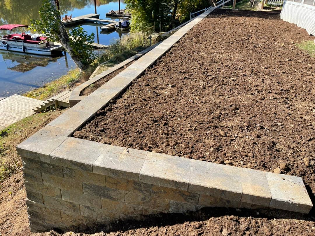 Landscape view of a freshly built stone retaining wall by a river with boats in the background.