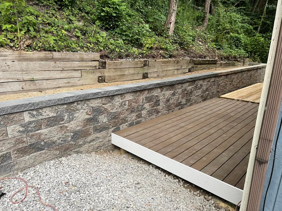 Landscape featuring a stone retaining wall and wooden deck in a wooded area.