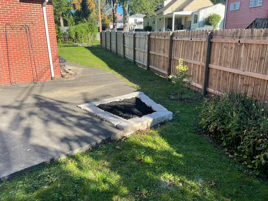 Backyard garden area with a soil bed surrounded by a brick path and wooden fence.