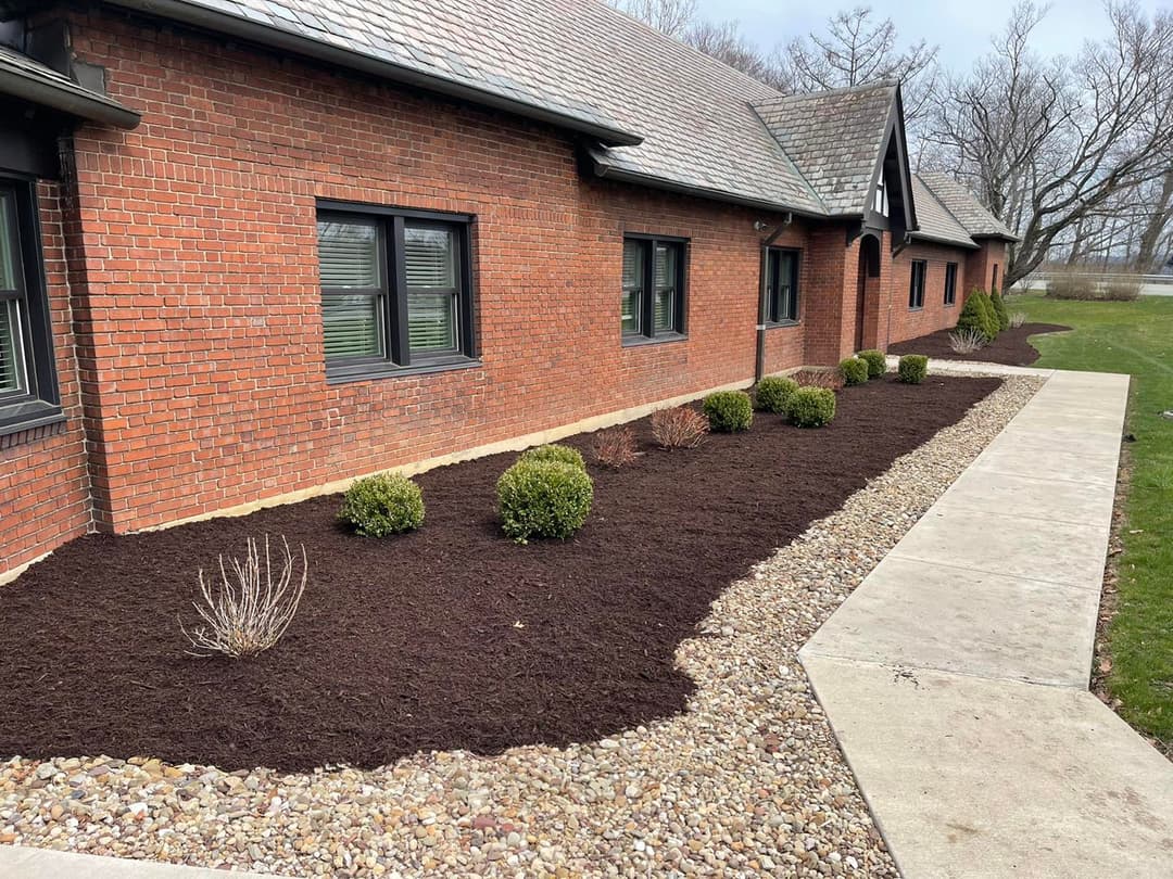 Mulched garden beds and neatly trimmed shrubs along a brick building's pathway.