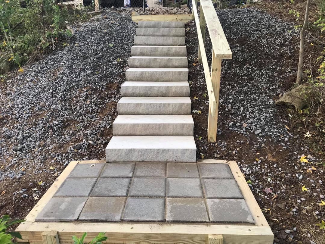 Concrete stairs with wooden railing leading up from gravel path in a landscaped area.
