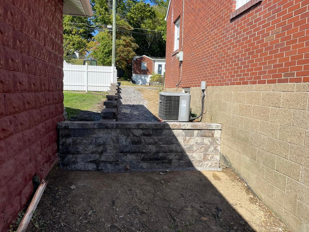 Stone retaining wall between two buildings with clear walkway and air conditioning unit.