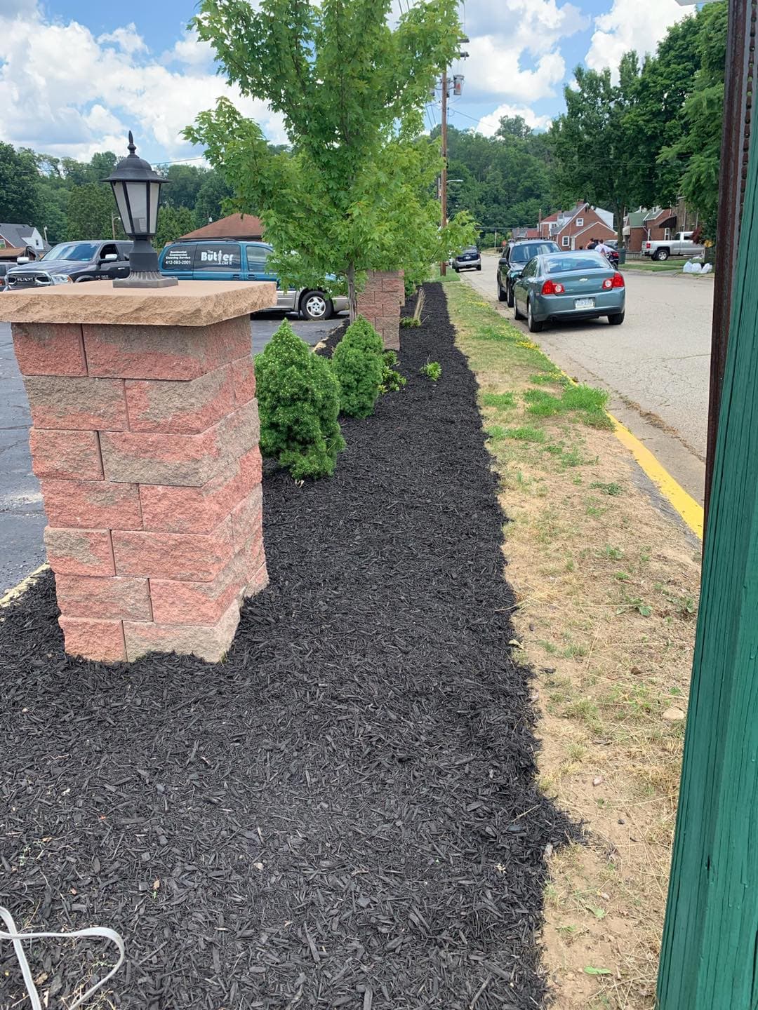 Landscaped area with mulch, bushes, and stone pillars beside a parking lot and trees.