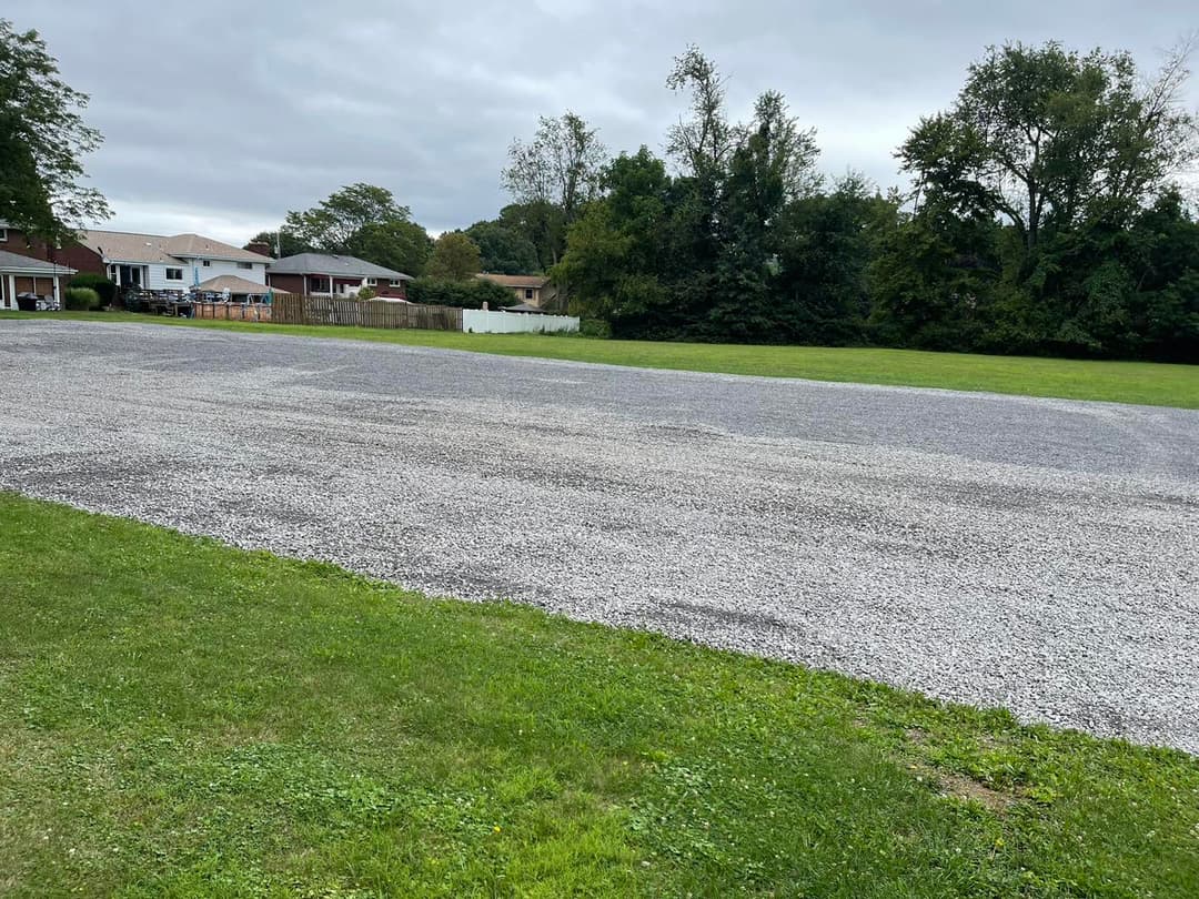 Gravel parking lot surrounded by green grass and suburban homes under a cloudy sky.