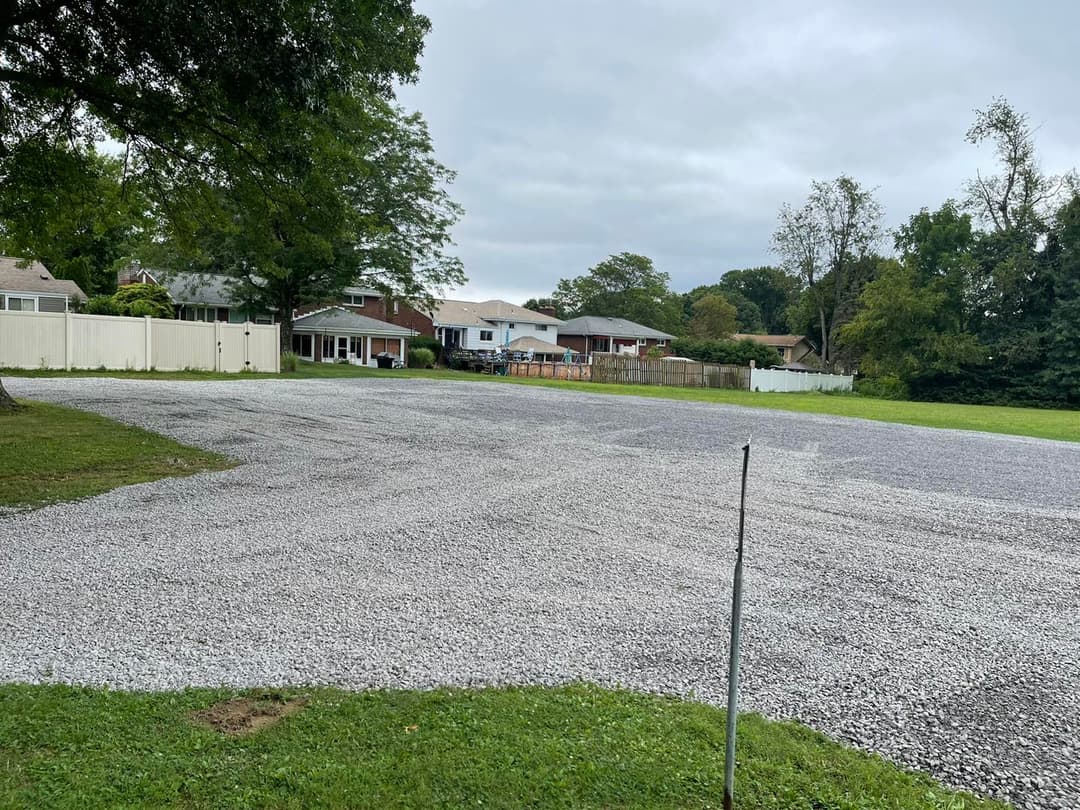 Gravel parking lot with homes and trees in the background under a cloudy sky.
