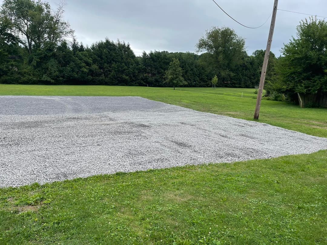 Gravel-covered area on a grassy field with trees and a power line in the background.