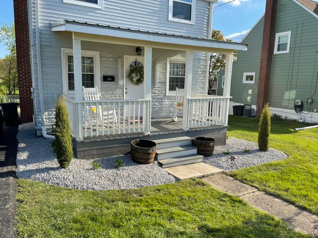 Front porch of a house with rocking chairs, potted plants, and a landscaped yard.