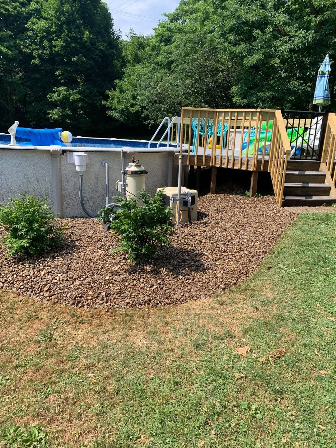 Backyard with a pool, wooden deck, landscaping, and pool equipment set in a sunny green area.