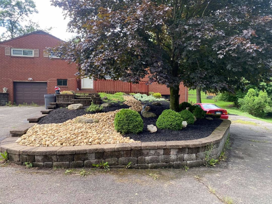 Landscape featuring stone garden beds, shrubs, and a large tree in a residential setting.