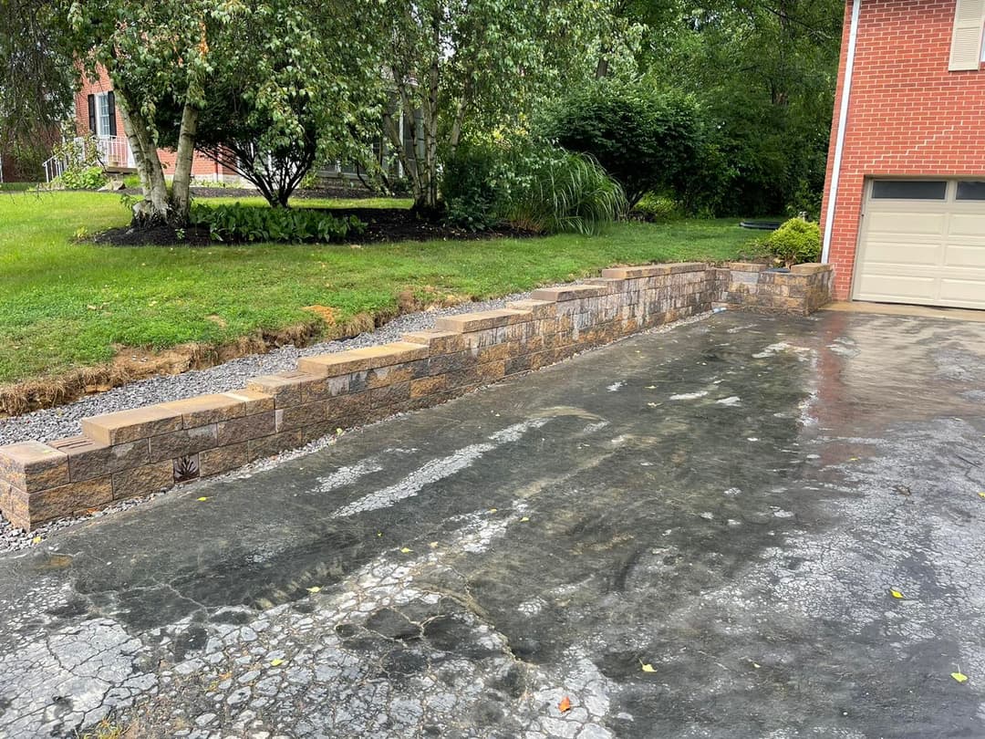 Stone retaining wall alongside a driveway, with grass and trees in the background.