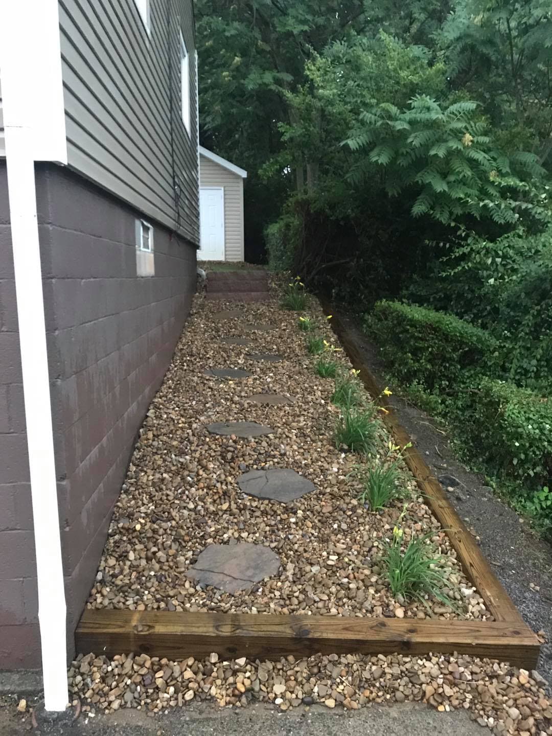 Rocky pathway lined with flowers alongside a house in a wooded area. Decorative landscaping.