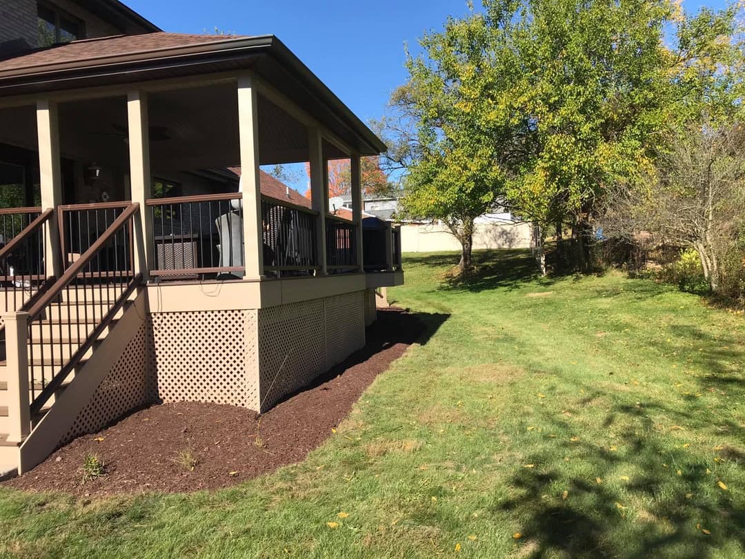 Backyard view of a house with a deck, green lawn, and trees under a clear blue sky.