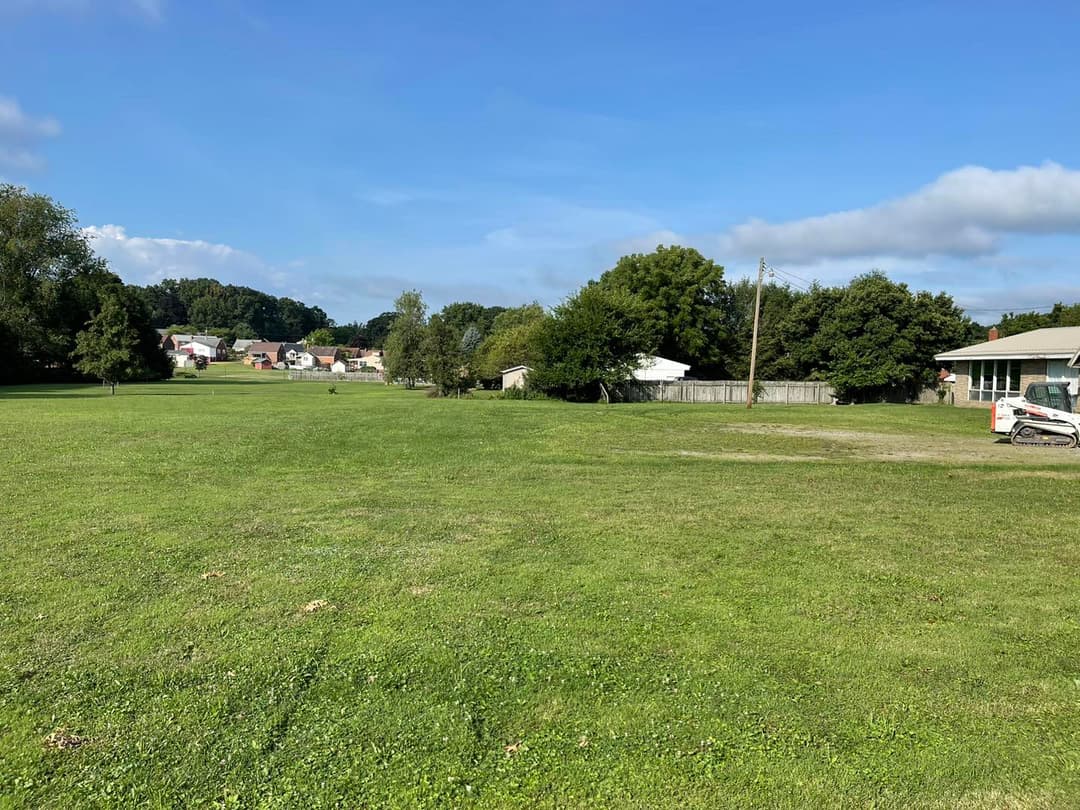 Open green field with trees and buildings in the background under a clear blue sky.