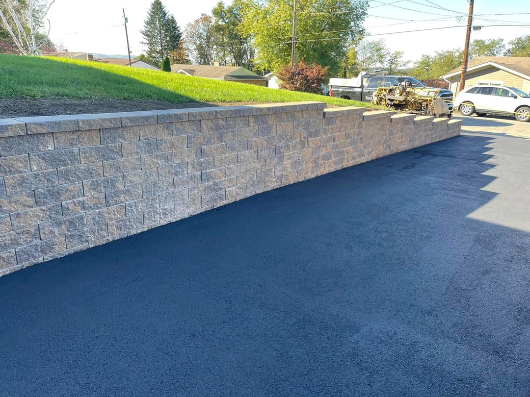 Retaining wall with stone blocks beside freshly paved black driveway and green lawn.