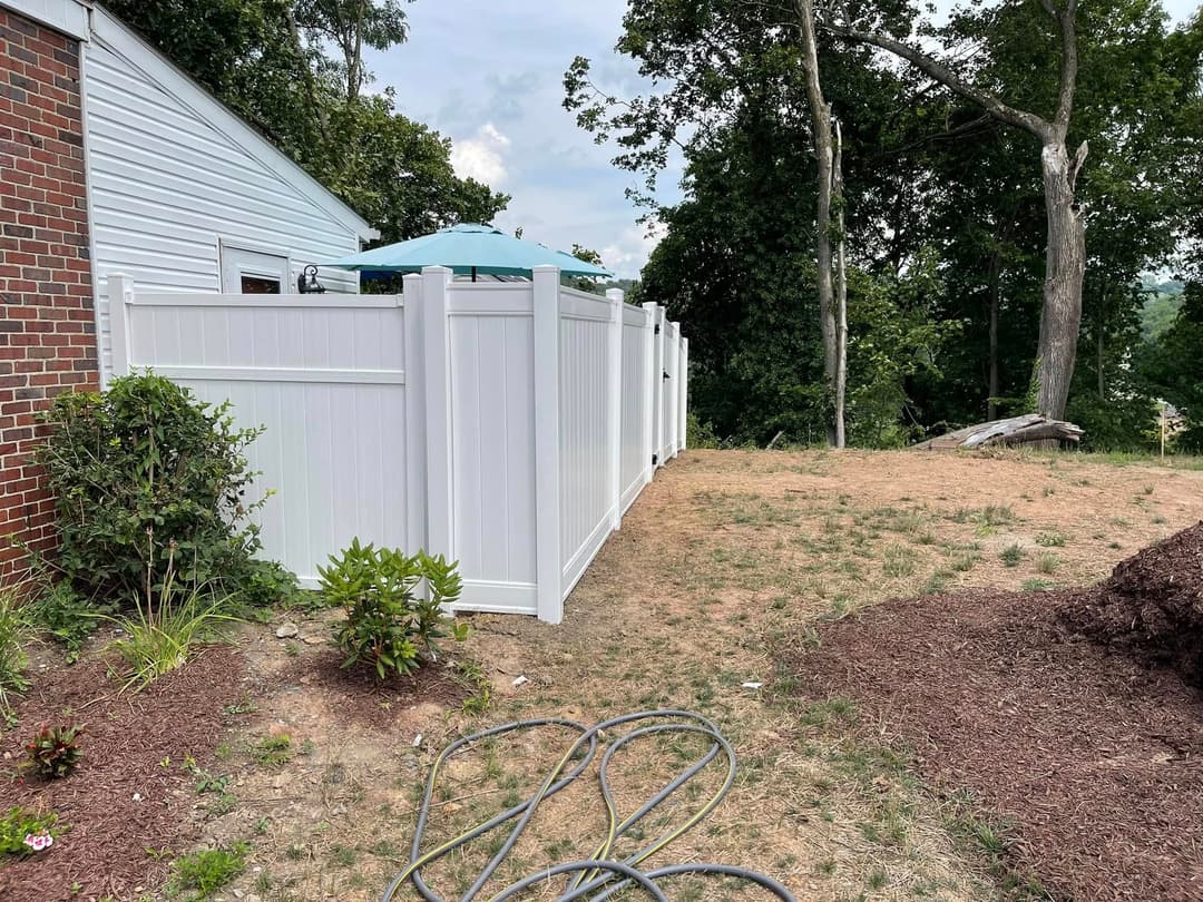 White vinyl fence surrounding a backyard area with grass and plants, featuring an umbrella shaded patio.