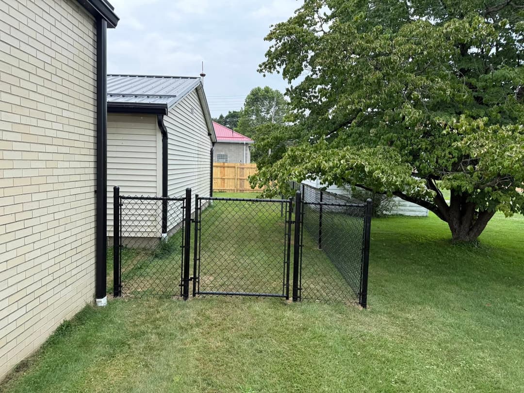 Chain link fence gate beside a house, leading to a grassy yard with a tree.