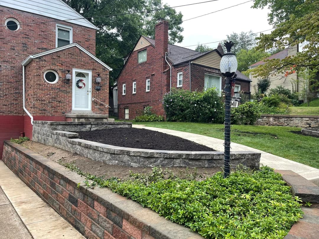 Front yard landscape featuring a stone garden bed, pathway, and brick wall near a residential home.