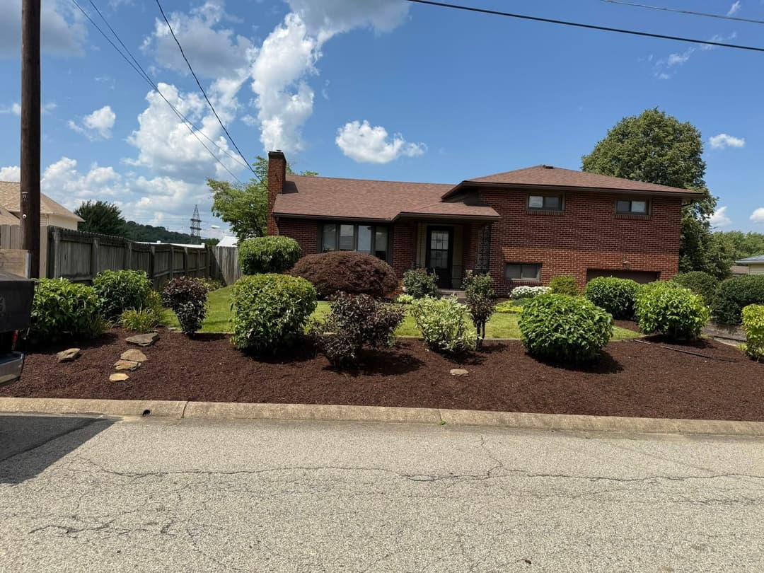Two-story brick house with manicured landscaping, green shrubs, and a clear blue sky.