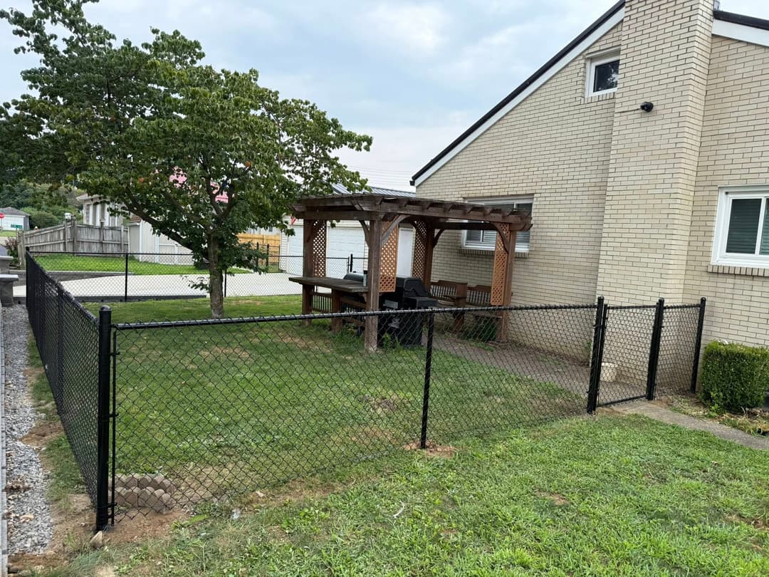 Chain-link fence enclosing a backyard patio with a pergola and seating area beside a brick house.